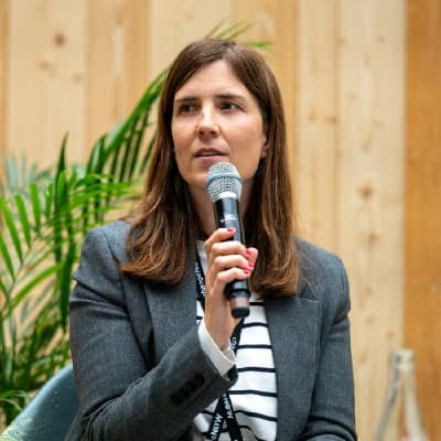 Frau im grauen Blazer spricht in ein Mikrofon vor einer Holzwand mit grüner Pflanze.