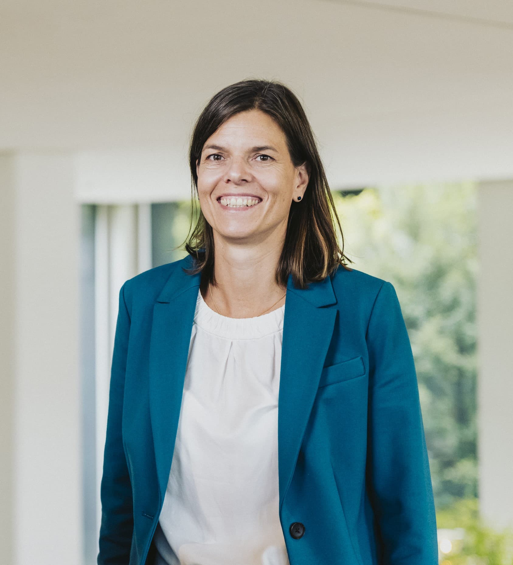 Lächelnde Frau mit braunen Haaren in einem blaugrünen Blazer und weißer Bluse vor hellem Hintergrund.