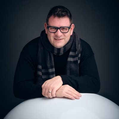 Ein lächelnder Mann mit Brille, schwarzem Pullover und gestreiftem Schal lehnt auf einer weißen Fläche.