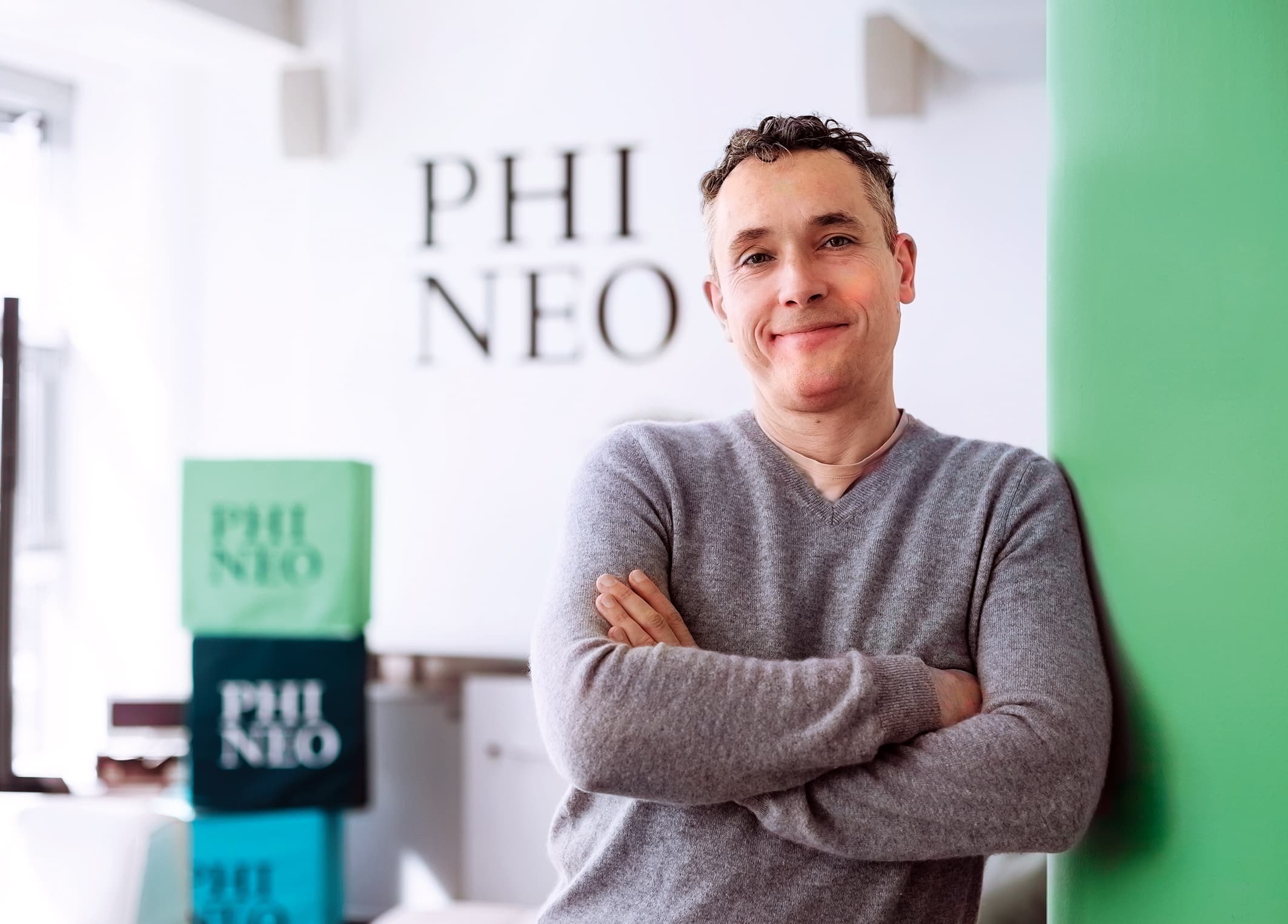 Lächelnder Mann mit verschränkten Armen lehnt an grüner Wand vor dem PHINEO-Logo im Hintergrund.