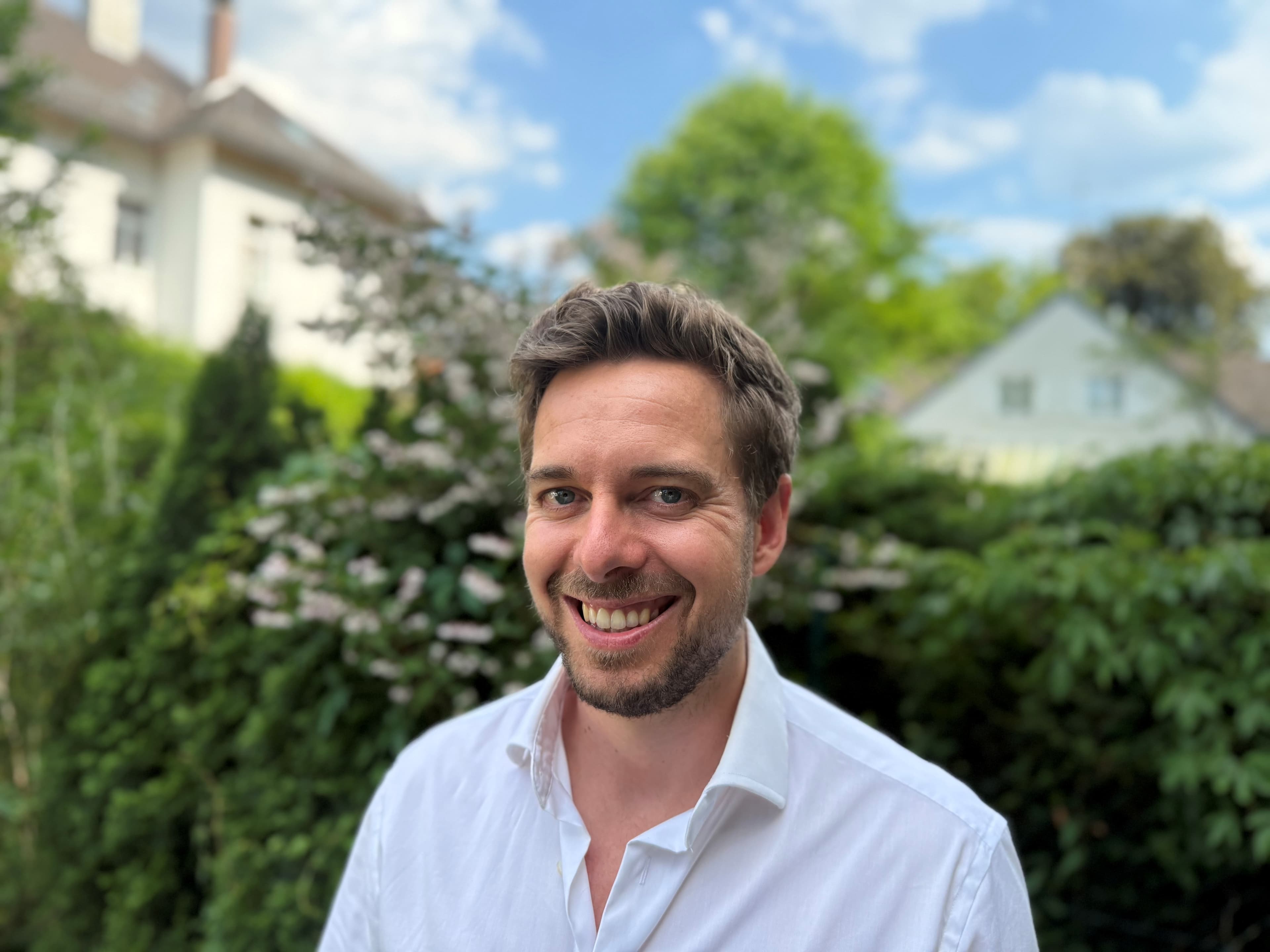Ein freundlich lächelnder Mann im weißen Hemd vor einem grünen Garten mit unscharfen Häusern.