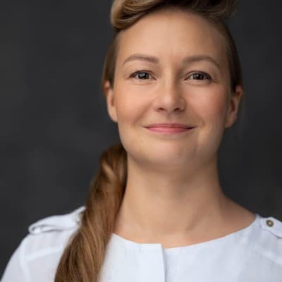 Porträt einer lächelnden Frau mit hellbraunem Haar, seitlichem Zopf und weißem Oberteil vor dunklem Hintergrund.