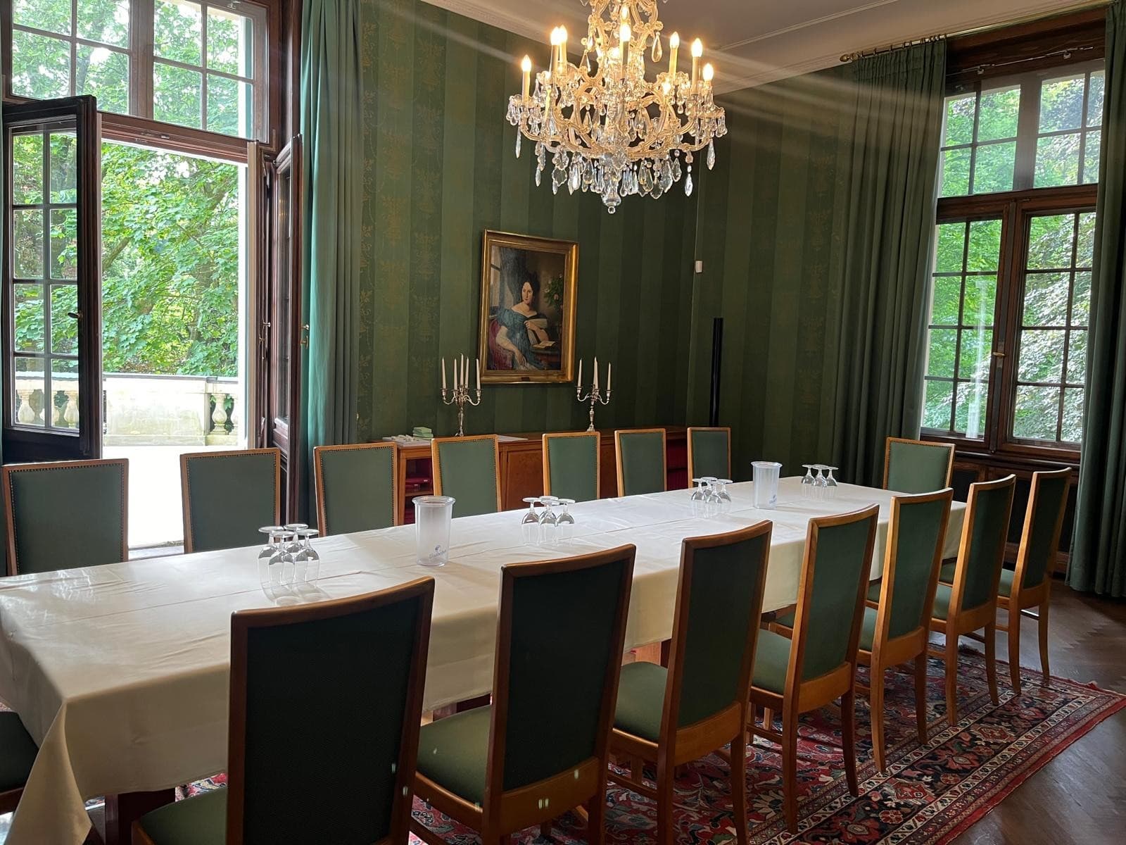 Eleganter Speisesaal mit langem weißem Tisch, grünen Stühlen, Kristallleuchter und klassischer grüner Tapete.