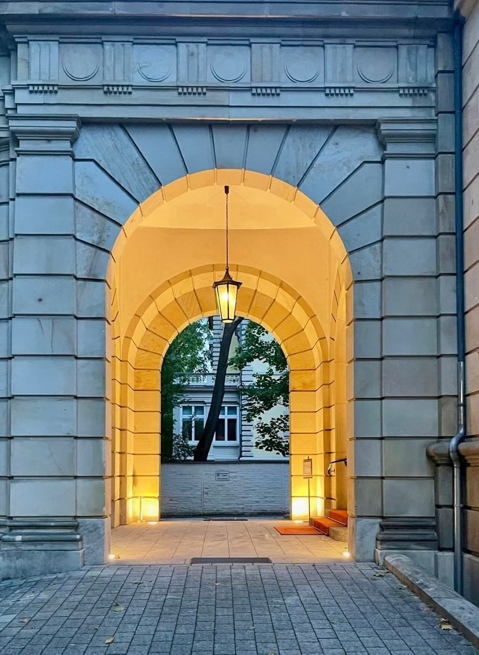 Ein klassischer Torbogen aus Stein mit hängender Laterne und warmem Licht führt in einen Innenhof.