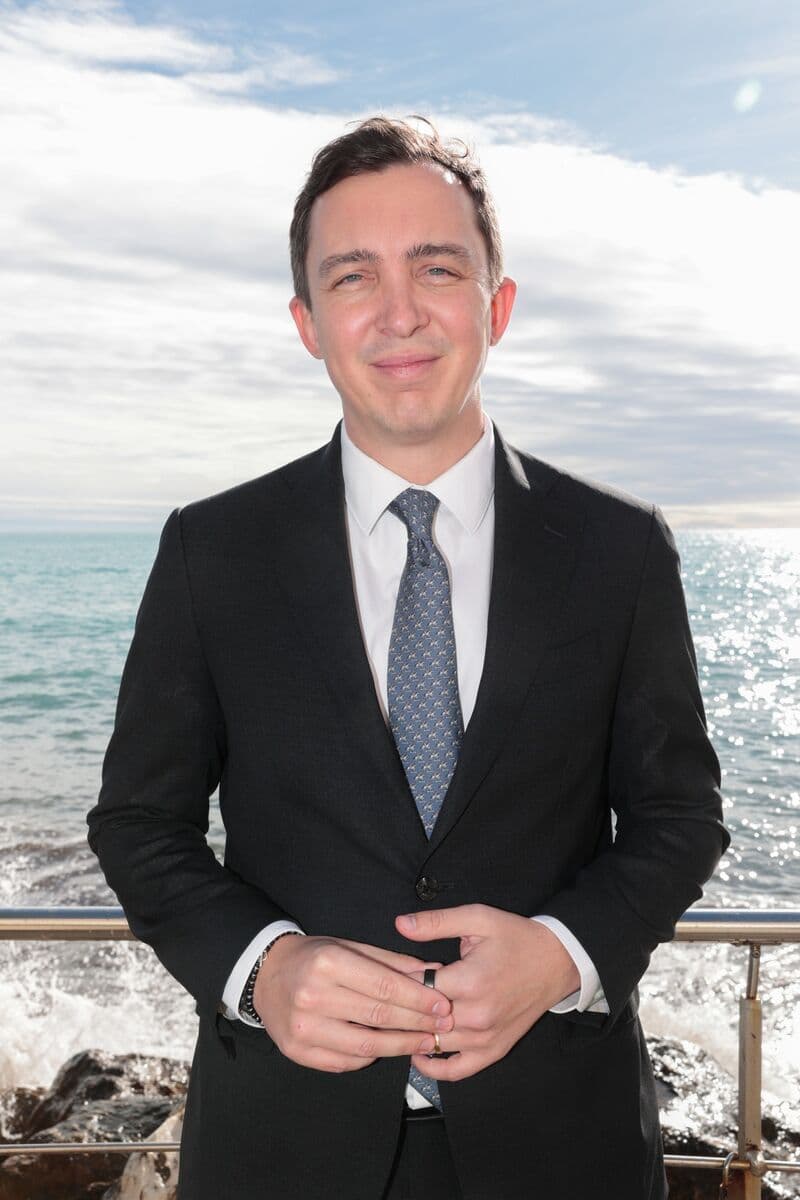 Ein lächelnder Mann im schwarzen Anzug und Krawatte vor dem glitzernden Meer unter bewölktem Himmel.